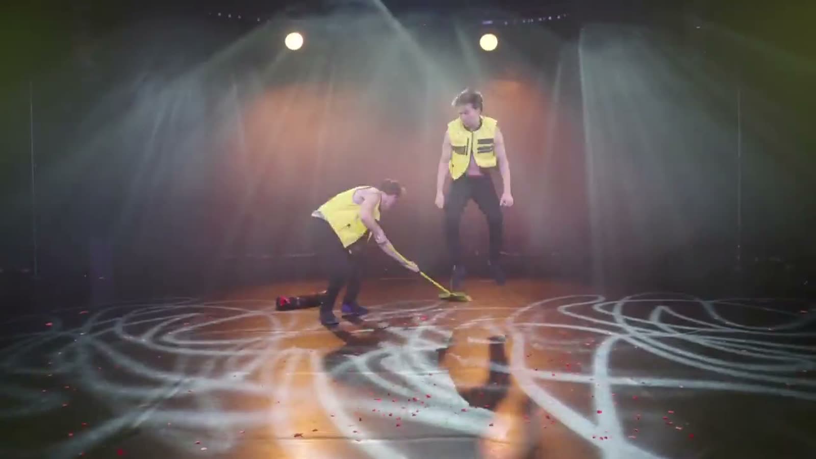 Jim & John in gelben Warnwesten auf einer leeren Bühne im Spotlicht, einer kehrt mit einem gelben Besen den Boden