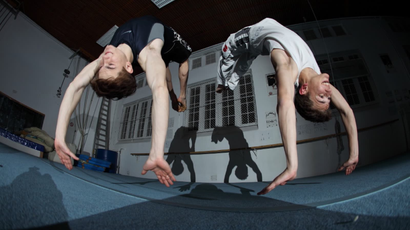 Beide Brüder gleichzeitig kopfüber im Rückwärtssalto in der Trainingshalle, ihre Schatten an der Wand dahinter